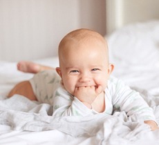 A baby lying on its stomach and grinning while chewing on its fingers before seeing its pediatric dentist in Garland